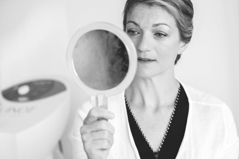 Une image en niveaux de gris d'une femme tenant un miroir à main et le regardant attentivement. Elle a une expression calme, porte un cardigan de couleur claire sur un haut à pois et a les cheveux tirés en arrière. L'arrière-plan présente une machine floue, indiquant peut-être un cadre esthétique pour les préparatifs de sa surprise de mariage.