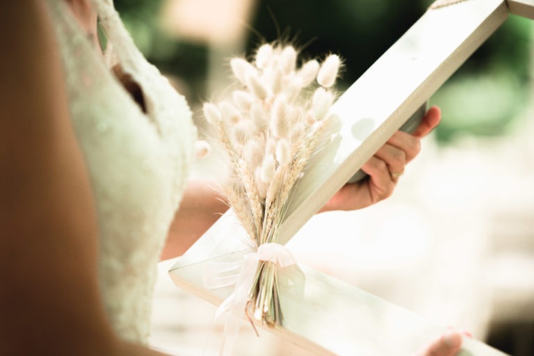 Gros plan d'une personne vêtue d'une robe blanche en dentelle tenant un cadre géométrique en bois. Le cadre présente un bouquet de fleurs séchées, notamment des tiges de blé et des têtes de graines duveteuses, attachées avec un ruban rose transparent. L'arrière-plan est flou, suggérant un décor extérieur avec de la verdure, un moment parfait pour une surprise de mariage.