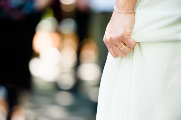 Gros plan sur la main d'une personne tenant délicatement le tissu d'une robe blanche, avec un bracelet délicat au poignet et une bague en or au doigt. L'arrière-plan est flouté par des lumières bokeh circulaires, suggérant un décor extérieur pour une surprise de mariage, avec la lumière du soleil filtrant à travers.