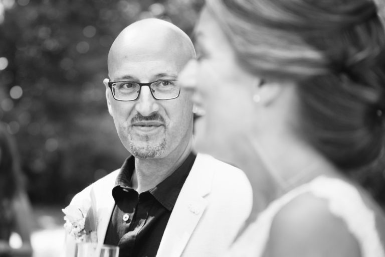 Photographie en noir et blanc d'un homme avec des lunettes, une tête chauve et une barbiche, vêtu d'un blazer clair et d'une chemise sombre, regardant attentivement une femme au premier plan. La femme, floue, a les cheveux longs coiffés en arrière. L'arrière-plan est flou avec des arbres, laissant deviner un décor extérieur pour une surprise de mariage.