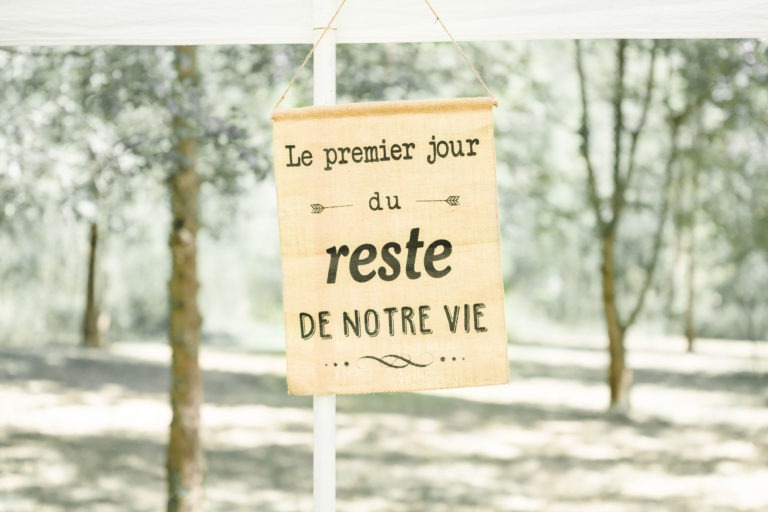 Une pancarte en toile de jute suspendue sous un auvent blanc porte l'inscription « Le premier jour du reste de notre vie », faisant allusion à une surprise de mariage. L'arrière-plan montre des arbres flous et la lumière du soleil filtrant à travers, créant un cadre extérieur serein.