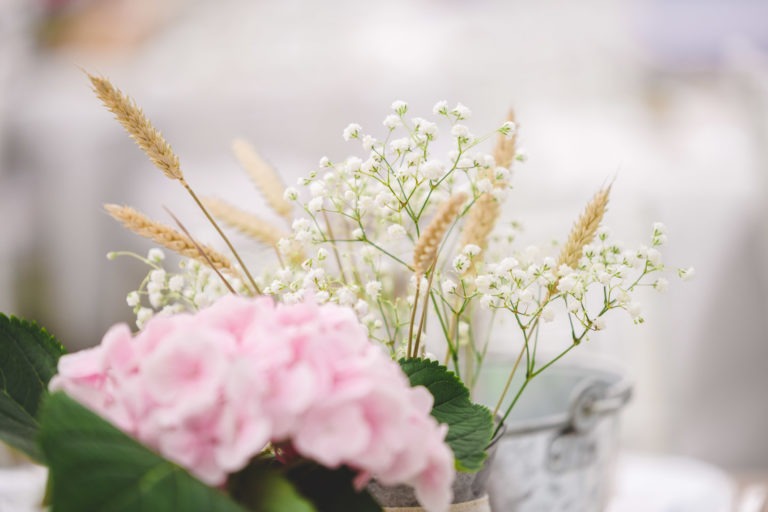 Une composition florale délicate composée d'hortensias rose pâle, de gypsophiles blancs et de tiges de blé beiges placées dans un vase gris clair. Parfait pour une surprise de mariage, l'arrière-plan est légèrement flouté, créant une ambiance sereine et élégante.