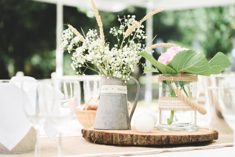 Un centre de table rustique comprend un pichet en métal rempli de gypsophile blanc et de tiges de blé, ainsi qu'un bocal en verre contenant une rose rose et des feuilles vertes. Tous deux sont placés sur une dalle de bois ronde, entourés de verrerie et de serviettes blanches, rappelant une élégante surprise de mariage sur fond vert extérieur flou.