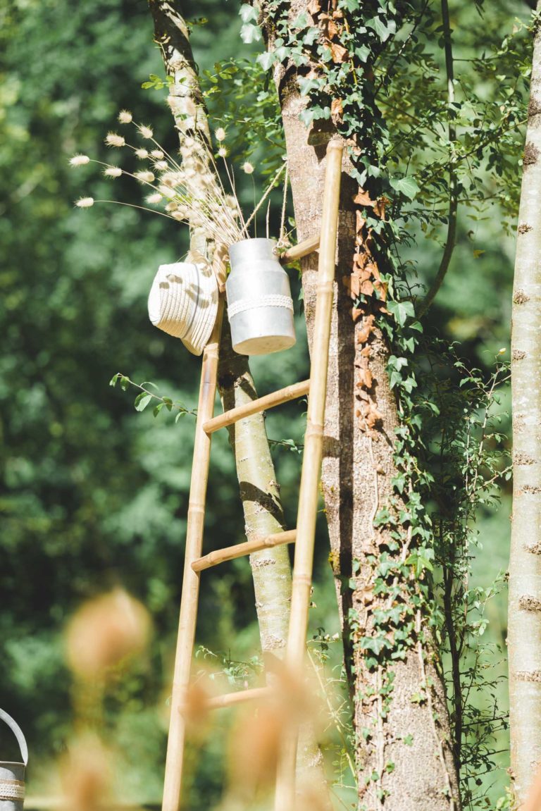 Une échelle en bois rustique est adossée à un arbre recouvert de lierre. Sur l'une des marches de l'échelle, il y a un seau en métal blanc rempli de fleurs séchées et un chapeau de paille est suspendu à côté. L'arrière-plan est rempli de verdure luxuriante, créant le cadre parfait pour une surprise de mariage intime.