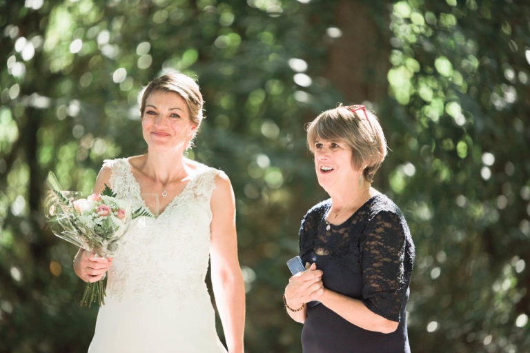 Une mariée en robe de dentelle blanche tient un petit bouquet de fleurs, debout à l'extérieur à côté d'une femme plus âgée en robe de dentelle bleu marine. Toutes deux sourient, avec de la verdure et des rayons de soleil créant un arrière-plan tacheté. La femme plus âgée, capturant ce moment spécial sur son smartphone, semble ravie de la surprise du mariage.