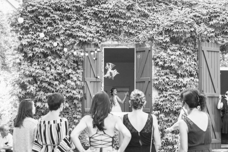 Une mariée se tient devant une porte ouverte encadrée par des murs couverts de lierre, se préparant à lancer son bouquet. Cinq femmes, vues de dos, attendent avec impatience de le récupérer. L'image en noir et blanc capture l'anticipation et la joie du moment. Les décorations rustiques ajoutent du charme à ce décor de mariage surprise.