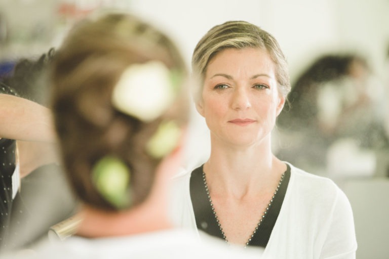 Une femme aux cheveux blonds, coiffée en chignon orné de fleurs blanches, se regarde dans un miroir. Elle porte un haut ou une robe de couleur claire et a une expression calme. Dans l'arrière-plan flou, une personne s'occupe de ses cheveux en prévision de ce qui semble être une surprise de mariage. Le décor semble être celui d'un salon ou d'un environnement similaire.