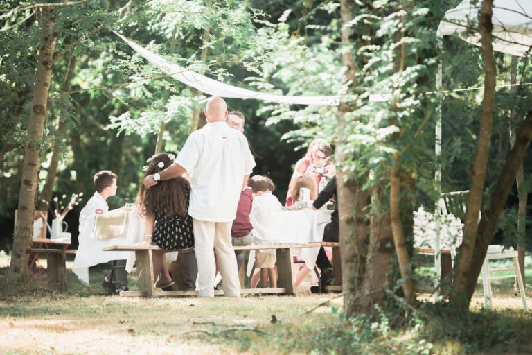 Un groupe de personnes se rassemble à l'extérieur pour profiter d'un pique-nique sous un soleil tamisé. Ils sont assis à une table ornée d'une nappe blanche, entourée de verdure luxuriante et d'arbres. Un homme en chemise blanche embrasse un enfant portant une couronne de fleurs. L'atmosphère est détendue et festive, évoquant l'atmosphère d'une surprise de mariage.