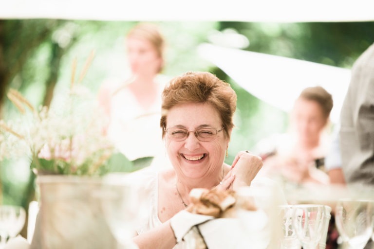 Une femme âgée aux cheveux châtains courts et aux lunettes sourit chaleureusement à une table dressée pour un repas en plein air. Elle est entourée de personnes aux contours flous et de verdure en arrière-plan, créant une ambiance lumineuse et joyeuse rappelant une célébration de mariage surprise. La table est ornée de verres, de fleurs et de pain.