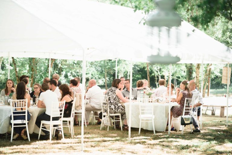 Un groupe de personnes est assis autour de tables rondes sous un grand auvent blanc lors d'un événement en plein air. Les tables sont drapées de nappes blanches et des chaises blanches sont disposées autour d'elles. Des arbres et de la verdure entourent la zone, créant un décor naturel et serein, parfait pour une surprise de mariage. Certaines personnes sont engagées dans une conversation.