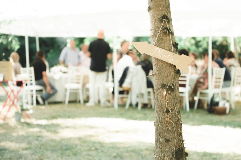 Au premier plan, un arbre avec un petit panneau en forme de flèche sur lequel est écrit « Photos » en lettres cursives. À l'arrière-plan, un groupe de personnes est assis autour de tables recouvertes de nappes blanches sous un auvent, en train de discuter lors d'un événement en plein air. La scène, préparée pour une surprise de mariage, se déroule dans une zone herbeuse avec lumière naturelle.