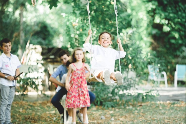 Un enfant joyeux, vêtu d'une chemise blanche et d'un short, se balance sur une balançoire, le sourire aux lèvres. À proximité, une fille en robe rose et un garçon en chemise bleue, assis, observent comme s'ils attendaient une agréable surprise. Un autre enfant se tient à gauche, tenant la corde de la balançoire. L'arrière-plan est un espace extérieur avec des arbres et du feuillage.