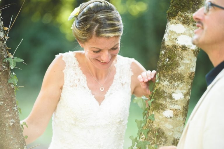 Une mariée en robe de mariée en dentelle blanche sourit et touche un tronc d'arbre orné de verdure. Elle porte une fleur dans les cheveux et un collier, incarnant l'essence d'une surprise de mariage. Un homme en veste claire, partiellement visible, se tient debout à droite, regardant la mariée. L'arrière-plan est flouté par la verdure.