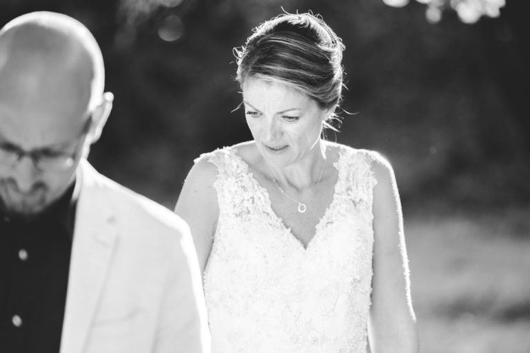 Une photo en noir et blanc montre une mariée dans une robe de mariée en dentelle avec un décolleté en V et des détails complexes, l'air pensif avec un soupçon de sourire. Ses cheveux sont relevés et elle porte un collier délicat. Au premier plan, flou, on voit un homme chauve portant des lunettes et un blazer de couleur claire, inconscient de la surprise du mariage qui l'attend.