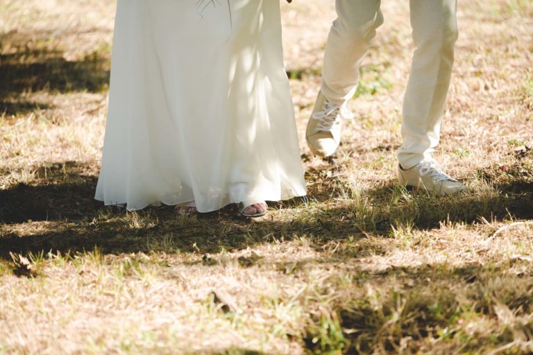 Gros plan d'un couple marchant en extérieur sur une pelouse ensoleillée. L'individu à gauche porte une robe blanche fluide, et celui à droite est vêtu d'un pantalon clair et de baskets blanches. Seuls leurs jambes et leurs pieds sont visibles, évoquant la spontanéité d'une promenade de mariage surprise. Les ombres des arbres parsèment le sol.