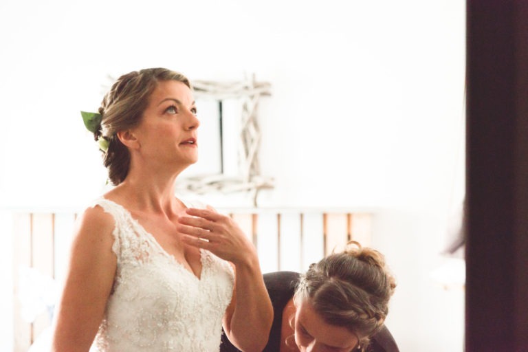 Une mariée en robe de mariée en dentelle blanche regarde vers le haut, la main droite près de sa poitrine. Elle a des fleurs dans ses cheveux tressés et se tient debout dans une pièce doucement éclairée. Un accompagnateur, qui fait partie de la surprise du mariage, examine la partie inférieure de sa robe. Un cadre en bois rustique est flou à l'arrière-plan.