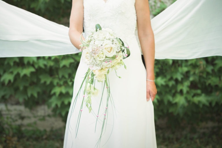 Une mariée en robe de mariée blanche tient un bouquet de fleurs blanches et crème avec un feuillage vert, sur fond de feuilles vertes et de tissu blanc drapé. L'accent est mis sur le bouquet et la moitié inférieure de la mariée, capturant l'essence de sa surprise de mariage.