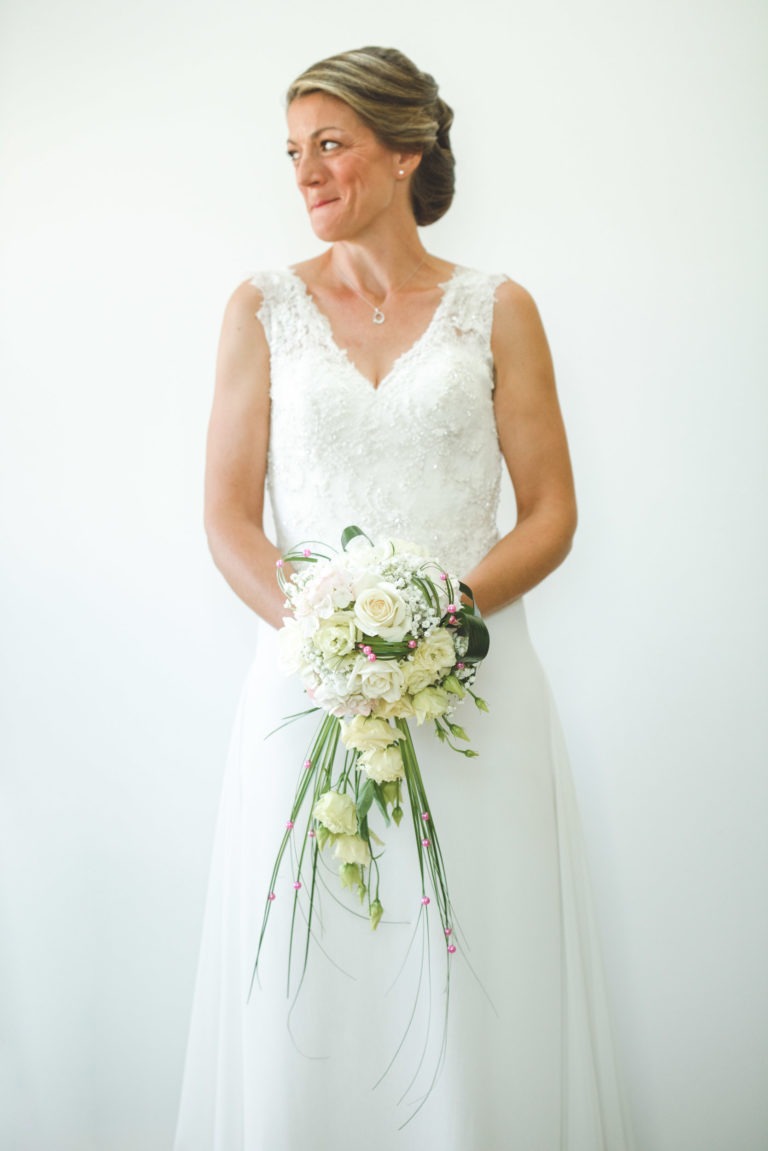 Une femme vêtue d'une robe de mariée sans manches en dentelle se tient debout sur un fond blanc uni, tenant un bouquet en cascade de fleurs blanches et de verdure. Elle a une coiffure relevée complexe et regarde vers sa gauche avec une expression sereine, comme si elle anticipait une surprise de mariage. Elle porte un collier pendentif et aucune boucle d'oreille visible.