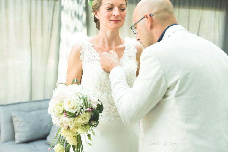 Une mariée en robe blanche tient un bouquet de fleurs blanches. Elle regarde vers l'avant tandis qu'un homme chauve portant des lunettes, un costume blanc et une chemise bleue, ajuste son collier. Ils sont à l'intérieur avec des rideaux gris et un canapé gris en arrière-plan, la lumière filtrant à travers. Serait-ce une surprise de mariage ?