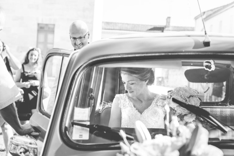 Une photo en noir et blanc montre une mariée assise dans une voiture ancienne, regardant vers le bas et souriant. Sa robe de mariée est finement conçue avec de la dentelle. En arrière-plan, un homme, peut-être le marié, et un autre individu se tiennent près de la porte ouverte. La scène de ce mariage surprise se déroule à l'extérieur par une journée ensoleillée.