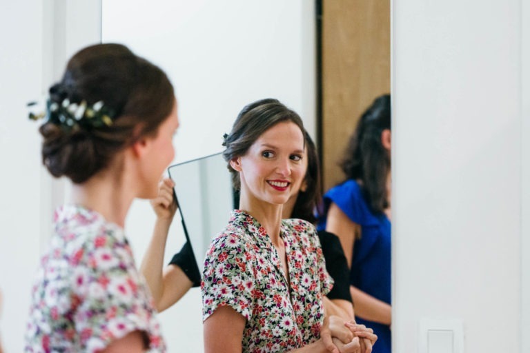 Une femme vêtue d'une robe fleurie se tient devant un miroir, souriant et admirant son reflet. Ses cheveux sont coiffés en un chignon élégant orné de petites fleurs. Elle tient ses mains jointes, tandis qu'une personne vêtue d'une robe bleue, peut-être coordonnée au Mariage Parfait à Bordeaux, est partiellement visible derrière elle.