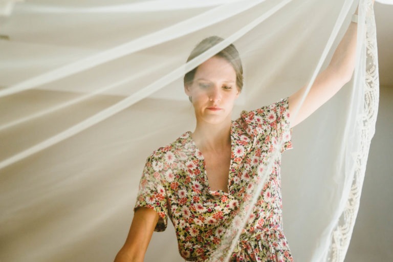 Une femme vêtue d'une tenue à motifs floraux, aux nuances de rouge et de rose, arrange soigneusement un tissu blanc translucide. Elle regarde le tissu avec une expression concentrée tout en le tenant. L'arrière-plan, rappelant un Parfait à Bordeaux pour un mariage, est doucement éclairé, mettant en valeur la texture délicate du tissu et de sa tenue.