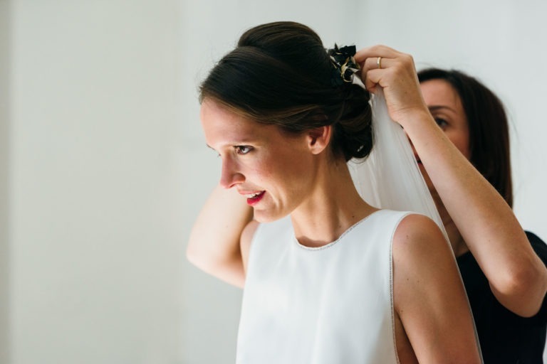 Une mariée en robe blanche sans manches sourit tandis qu'une autre femme ajuste ses cheveux, fixant un voile avec des épingles à cheveux. Les cheveux noirs de la mariée sont coiffés en chignon élégant, et une lumière illumine doucement son visage. Un mur uni et clair sert de fond à ce moment de mariage parfait à Bordeaux.