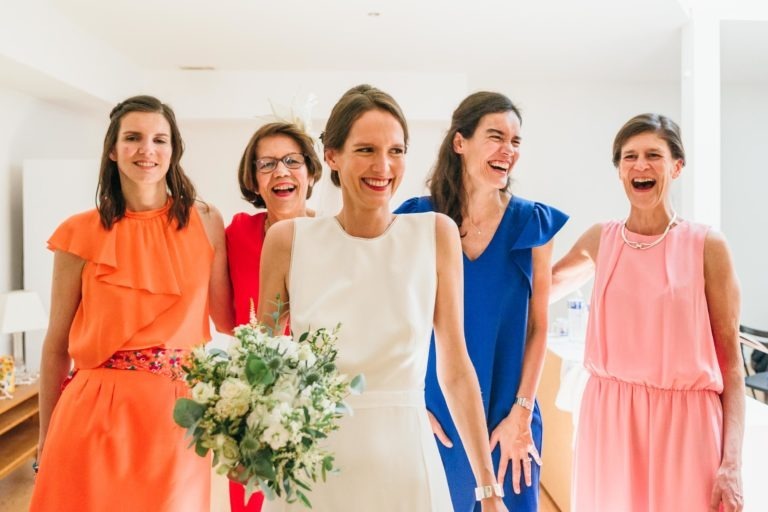 Une mariée en robe blanche tient un bouquet de fleurs blanches et de verdure, entourée de quatre femmes joyeuses en robes aux couleurs vives (orange, rouge, bleu et rose). Elles rient toutes et se tiennent debout dans un intérieur moderne et bien éclairé avec des murs blancs et un plancher en bois, une scène de mariage parfait à Bordeaux.