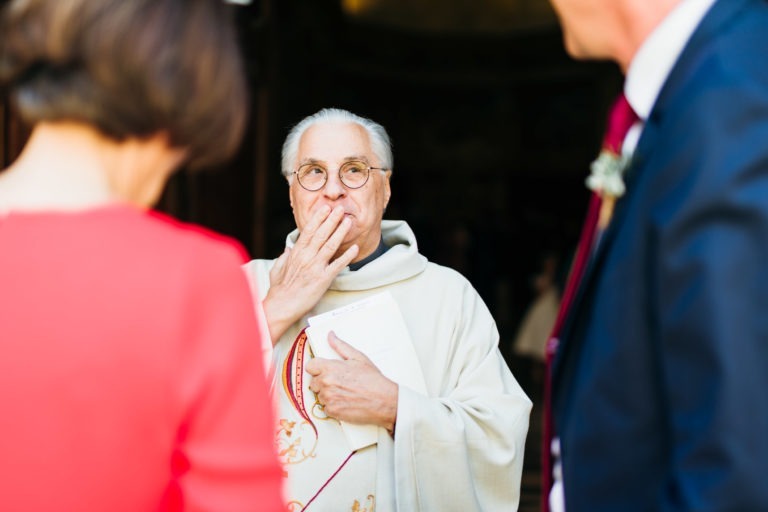 On voit un homme âgé en tenue religieuse et lunettes rondes tenant un document et portant la main à sa bouche dans un geste, peut-être pour célébrer un mariage parfait à Bordeaux. L'attention est portée sur lui, tandis que deux autres personnes, l'une vêtue de rouge et l'autre en costume, sont visibles mais floues au premier plan.