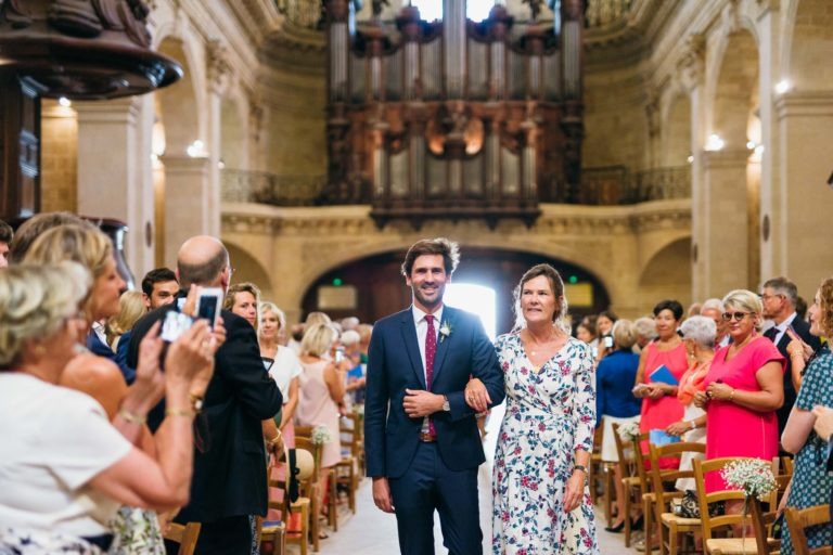 Un homme en costume bleu marine marche bras dessus bras dessous avec une femme en robe blanche à fleurs dans l'allée d'une grande église décorée. Les invités, habillés en tenue de soirée, se tiennent debout et regardent. L'arrière-plan met en valeur de hauts plafonds, une architecture complexe et un grand orgue à l'avant - un mariage parfait à Bordeaux.