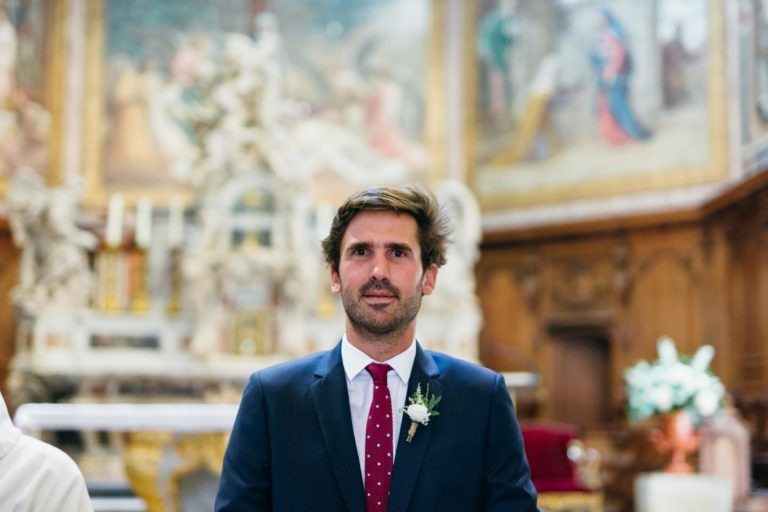 Un homme aux cheveux bruns et à la barbe se tient debout à l'intérieur, vêtu d'un costume bleu marine, d'une chemise blanche et d'une cravate à pois rouges. Il a une petite boutonnière épinglée au revers de son revers. L'arrière-plan présente un intérieur orné d'œuvres d'art détaillées, de boiseries et d'un autel - un véritable mariage parfait à Bordeaux.