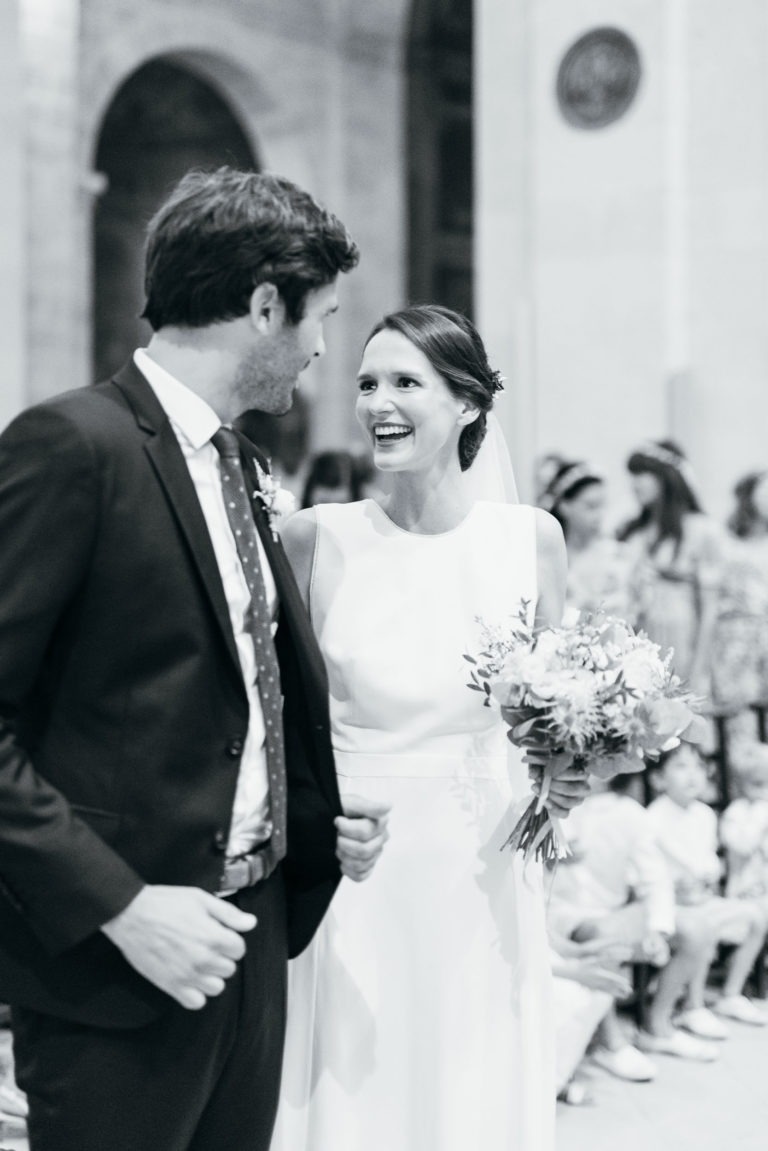 Un couple de mariés se tient debout dans un bâtiment et se sourit joyeusement. La mariée porte une robe blanche sans manches et tient un bouquet de fleurs. Le marié porte un costume sombre avec une cravate et une boutonnière. On peut voir plusieurs personnes à l'arrière-plan, assises et debout : mariage parfait à Bordeaux.