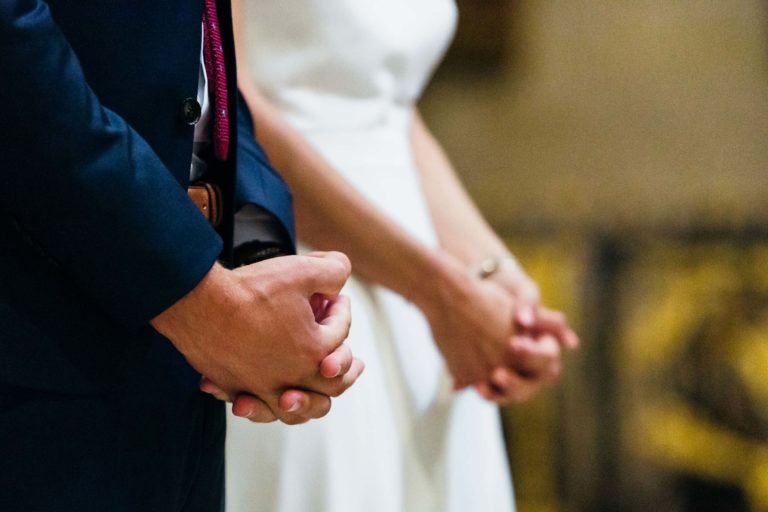 Deux personnes se tiennent côte à côte, les mains jointes devant elles. La personne de gauche porte un costume bleu foncé avec une cravate marron, celle de droite est en tenue blanche. Avec un arrière-plan flou mettant en valeur leurs mains et leur tenue, c'est comme si elles avaient trouvé leur mariage parfait à Bordeaux.