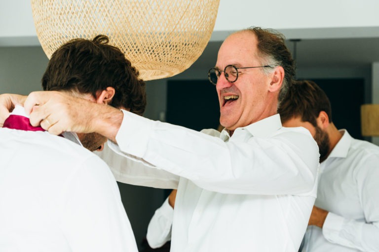 Un homme d'un certain âge portant des lunettes, souriant et ajustant le col de la chemise d'un homme plus jeune dans une pièce bien éclairée. Tous deux portent des chemises blanches et se préparent pour un mariage Parfait à Bordeaux. Un autre homme en chemise blanche est à l'arrière-plan, partiellement visible, apparemment occupé à une tâche similaire. Une lampe en osier est suspendue au-dessus.