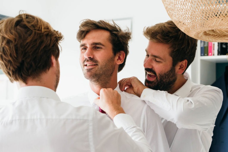 Deux hommes aident un autre homme à ajuster sa cravate. Tous trois sont vêtus de chemises blanches et se préparent pour un événement officiel au Mariage Parfait à Bordeaux. L'homme au milieu sourit et regarde sur le côté. Un luminaire tissé est partiellement visible dans le coin supérieur droit. Des livres et un costume sont à l'arrière-plan.