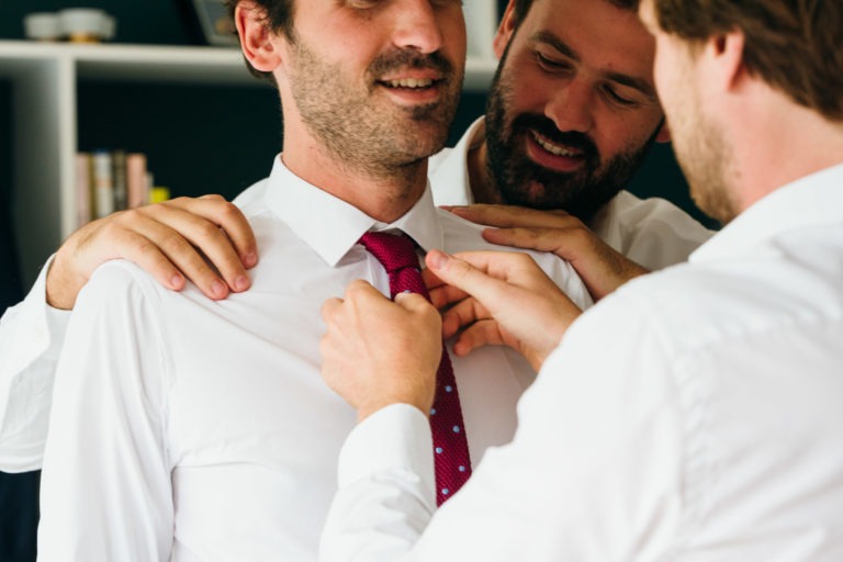 Trois hommes, vêtus de chemises blanches, s'entraident pour ajuster leurs cravates dans une pièce avec des étagères. L'homme du milieu, portant une cravate rouge, reçoit l'aide des autres tout en souriant. Cette scène de camaraderie laisse entrevoir les préparatifs d'un mariage parfait à Bordeaux.