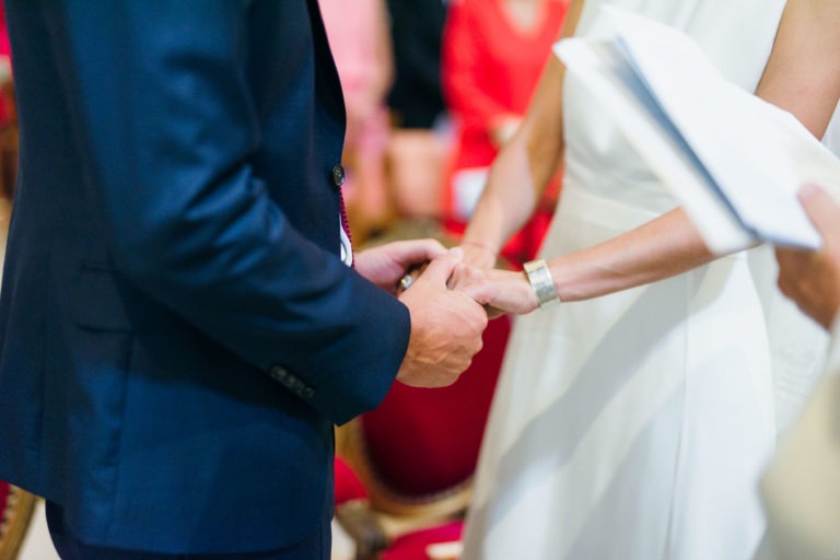 Une image en gros plan montre un couple se tenant la main lors d'un mariage parfait à Bordeaux. Le marié porte un costume bleu marine et la mariée est parée de sa robe blanche. Leurs visages ne sont pas visibles, tandis qu'une autre personne, partiellement visible à droite, tient un document, probablement l'officiant lisant les vœux.