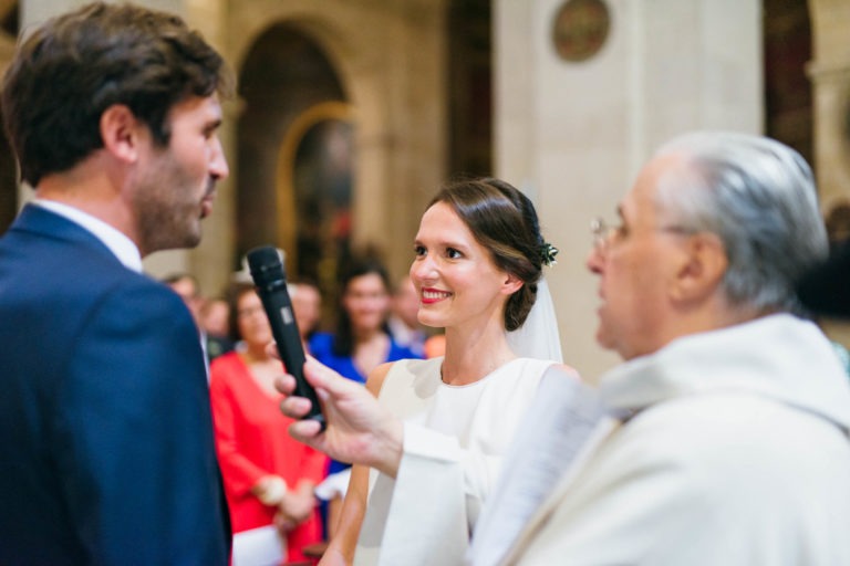 Un couple de mariés se tient devant l'autel lors de leur mariage Parfait à Bordeaux. La mariée, vêtue d'une robe blanche et d'un voile, sourit chaleureusement au marié, qui est habillé d'un costume bleu. Un prêtre en robe blanche leur tend un micro, avec d'autres invités flous en arrière-plan.