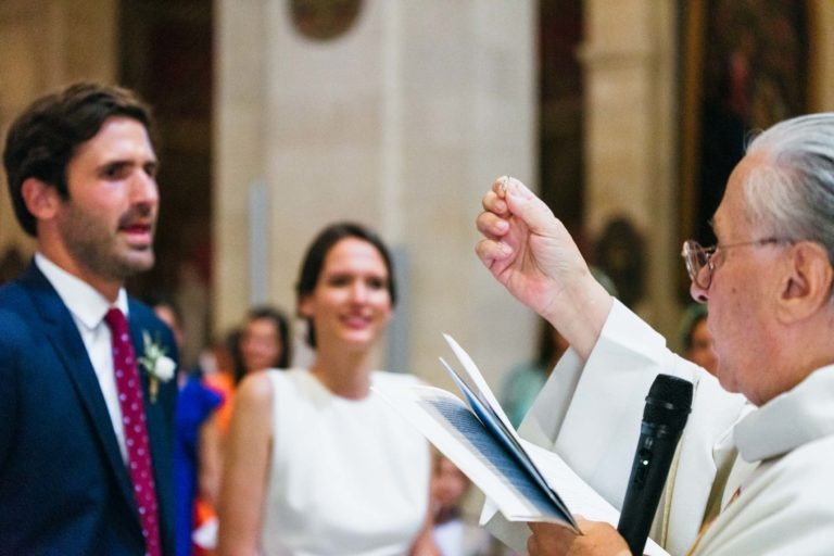 Un prêtre célèbre une cérémonie de mariage dans une église, tenant une bague devant lui, tandis que les mariés se tiennent devant lui. La mariée sourit et porte une robe blanche, tandis que le marié, vêtu d'un costume bleu avec une cravate à pois rouges, regarde. Ce mariage parfait à Bordeaux est observé par plusieurs invités flous en arrière-plan.