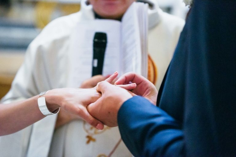 Gros plan d'une cérémonie de mariage Parfait à Bordeaux où un marié, vêtu d'un costume sombre, place une bague au doigt de la mariée. La main de la mariée est tendue, montrant un bracelet en argent. Un prêtre, tenant un livre et un microphone, est à l'arrière-plan, vêtu d'une robe cléricale blanche.