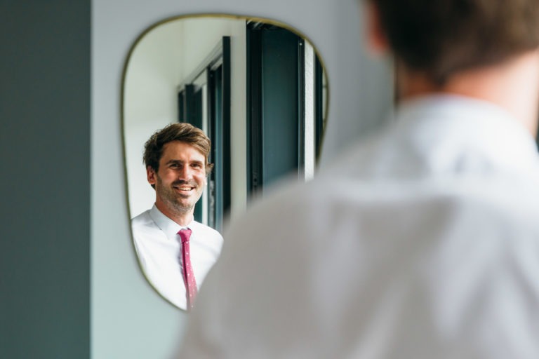 Un homme aux cheveux bruns et à la barbe courte sourit en regardant son reflet dans un miroir ovale. Il porte une chemise blanche et une cravate rouge à petits pois blancs, se préparant pour son Mariage Parfait à Bordeaux. La vue est par-dessus son épaule, capturant principalement son reflet, avec l'homme lui-même flou au premier plan.