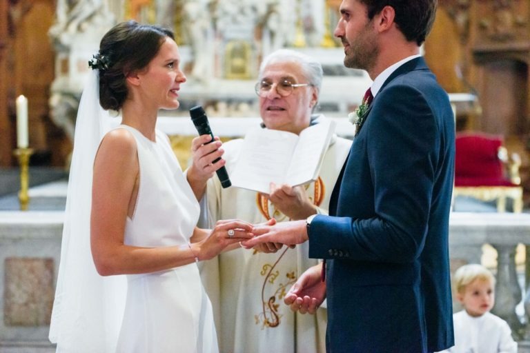 Un couple de mariés échange ses alliances lors de leur mariage Parfait à Bordeaux. La mariée, vêtue d'une robe blanche avec un voile, tient un micro et glisse une bague au doigt du marié. Le marié, vêtu d'un costume bleu, la regarde avec amour. Un prêtre en robe blanche se tient entre eux, tenant un livre ouvert.