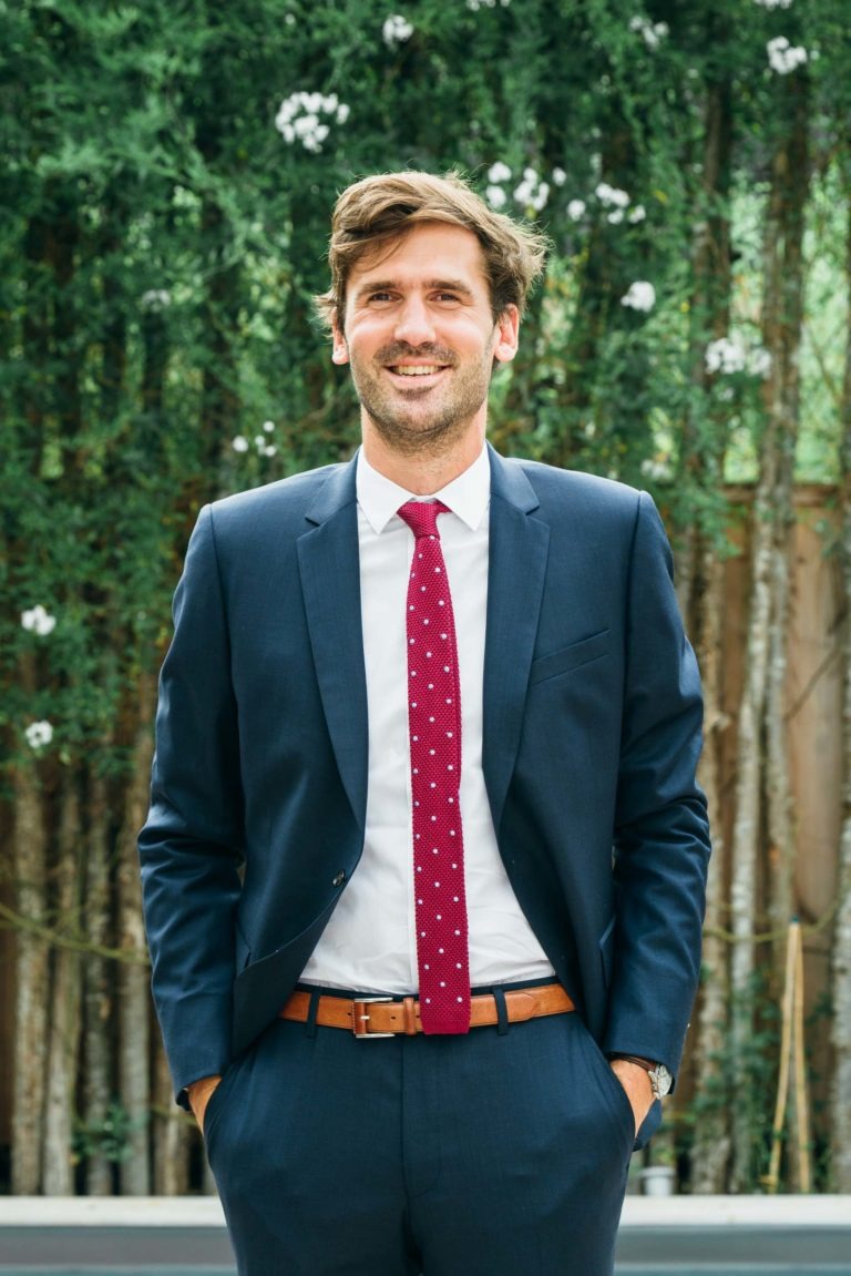 Un homme aux cheveux châtain clair et à la barbe courte se tient dehors devant des arbres verts. Il porte un costume bleu foncé, une chemise blanche et une cravate rouge à pois blancs. Les mains dans les poches et souriant, il semble prêt pour le mariage parfait Parfait à Bordeaux.