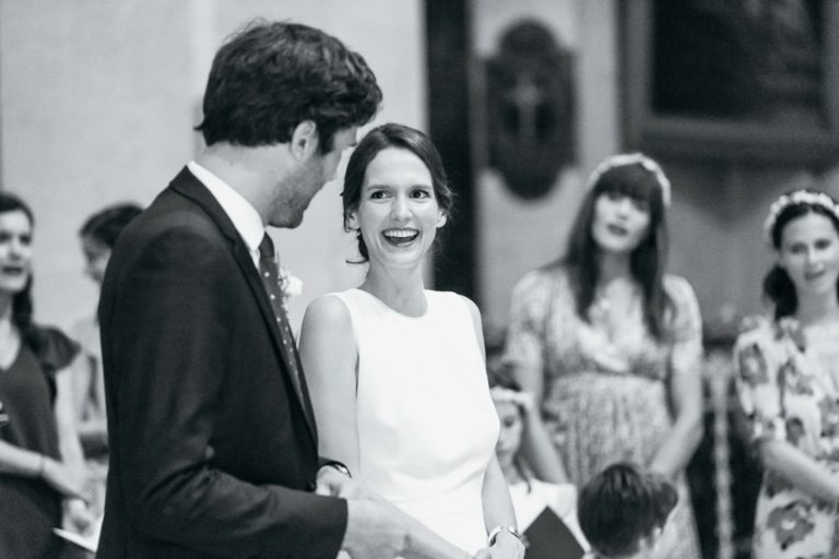 Une photo en noir et blanc montre une mariée souriante dans une robe blanche sans manches et un marié en costume sombre lors de leur mariage parfait à Bordeaux. Ils se tiennent face à face, la mariée regardant le marié. Les invités en robe se tiennent derrière eux, certains tenant des bouquets de fleurs.