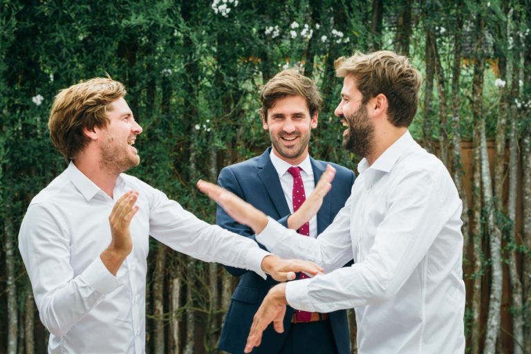 Trois hommes en plein air discutent avec animation. Deux d'entre eux, vêtus de chemises blanches, se font des gestes animés en souriant. Le troisième homme, vêtu d'un costume bleu marine et d'une cravate rouge, se tient entre eux, souriant également. La verdure forme le décor, suggérant une atmosphère décontractée et amicale rappelant un mariage parfait à Bordeaux.