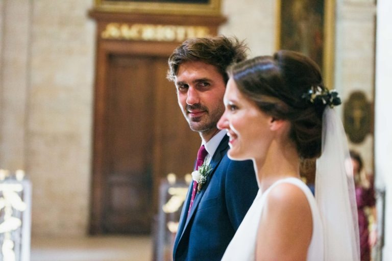 Un marié en costume bleu marine et cravate rouge avec une boutonnière sourit en regardant sa future épouse. La mariée, avec ses cheveux relevés en chignon orné d'un voile blanc, est vêtue d'une robe blanche. Ils se tiennent dans une église avec une porte en bois floue et des détails ornementaux en arrière-plan - un véritable mariage parfait à Bordeaux.