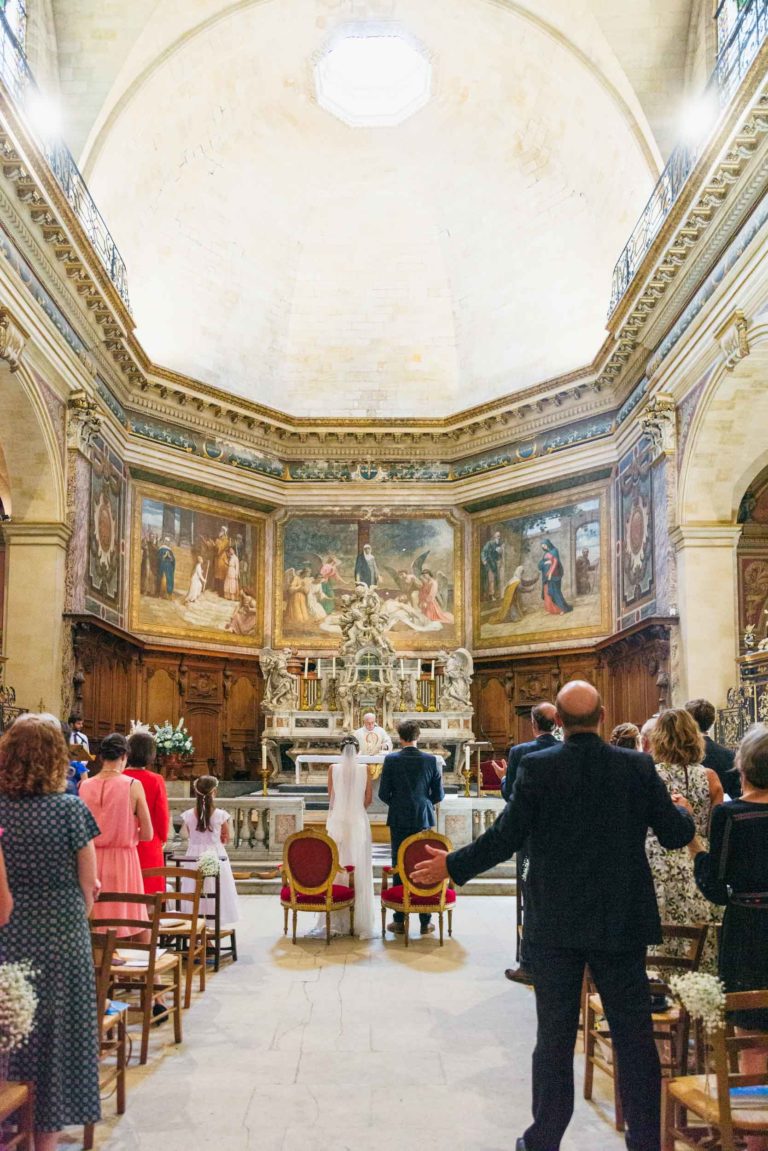 Un groupe de personnes se tient debout à l'intérieur d'une grande église de Bordeaux, face à l'autel. L'église a de hauts plafonds décorés, de grandes peintures sur les murs et des détails en bois complexes. Un prêtre se tient devant l'autel, qui comporte des statues et des décorations religieuses. La congrégation comprend des femmes en robe et des hommes en costume, célébrant un mariage parfait.