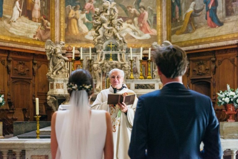 Un prêtre se tient devant l'autel, tenant un livre et faisant face aux mariés pendant leur mariage Parfait à Bordeaux. L'autel est décoré de bougies allumées, de fleurs et d'œuvres d'art religieuses complexes. La mariée, en robe blanche, et le marié, en costume sombre, se tiennent dos à la caméra.