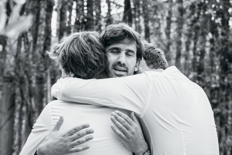 Trois hommes en chemise blanche se serrent dans leurs bras, l'air ému. L'homme au centre a les yeux fermés et semble en larmes. La photographie en noir et blanc capture un moment intime, rappelant un mariage parfait à Bordeaux, avec un arrière-plan boisé offrant un environnement naturel serein.