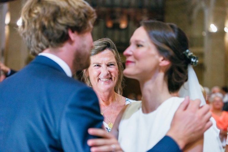 Une femme souriante se tient à l'arrière-plan, observant un couple le jour de leur mariage. Le marié, aux cheveux châtain clair, porte un costume bleu, tandis que la mariée, les cheveux attachés en arrière et vêtue d'une robe blanche, a les mains posées sur les épaules du marié. Le décor semble être celui d'une cérémonie en intérieur de mariage Parfait à Bordeaux.